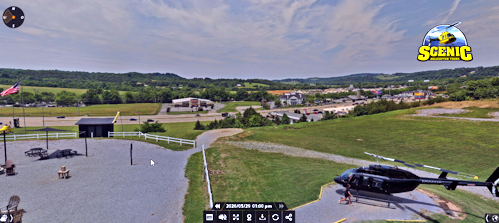 Aerial view of a park-like complex with a small helicopter on a helipad to the right, gravel paths, white fences, and green fields; SCENIC logo visible in the top-right corner.